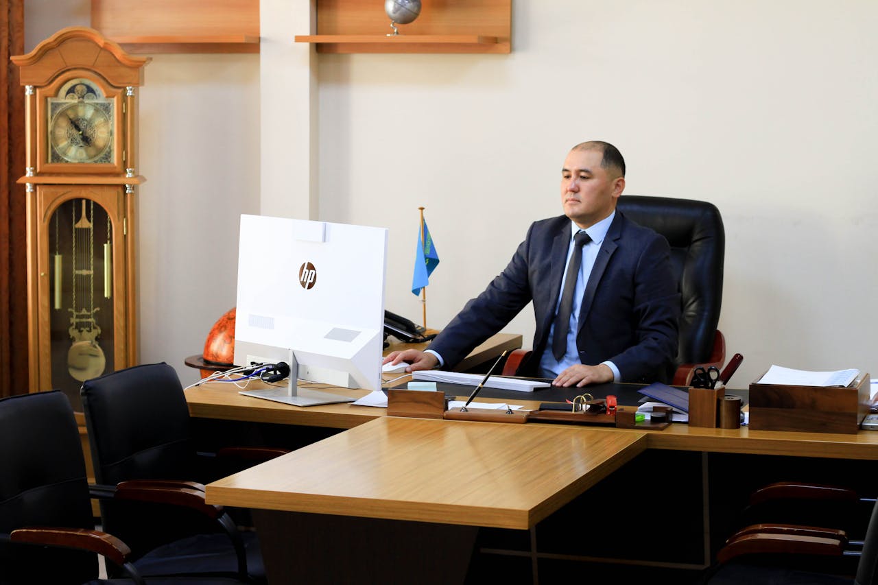 A serious businessman in a black suit focuses on work at his office desk with a computer and office decor.