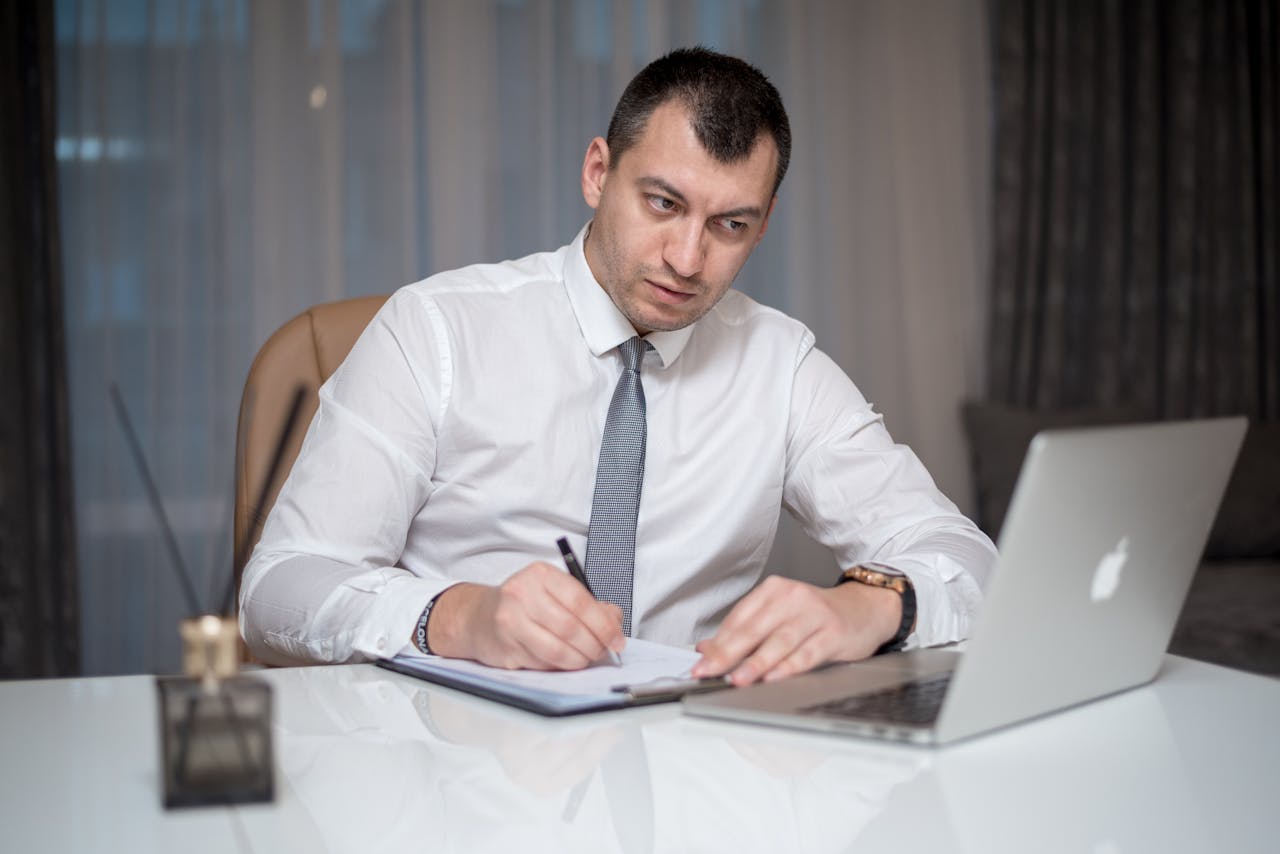 Focused businessman in white shirt writing notes at desk with laptop.