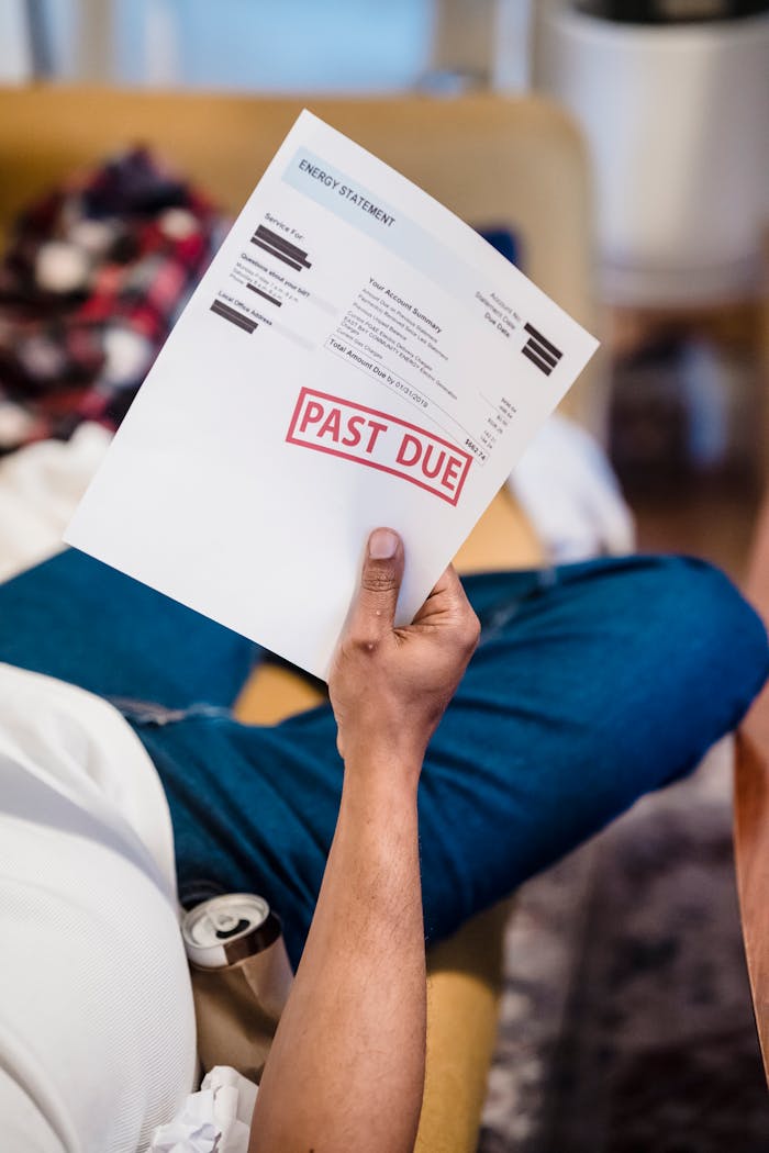 A man holding a past due billing statement while lounging indoors, expressing financial stress.